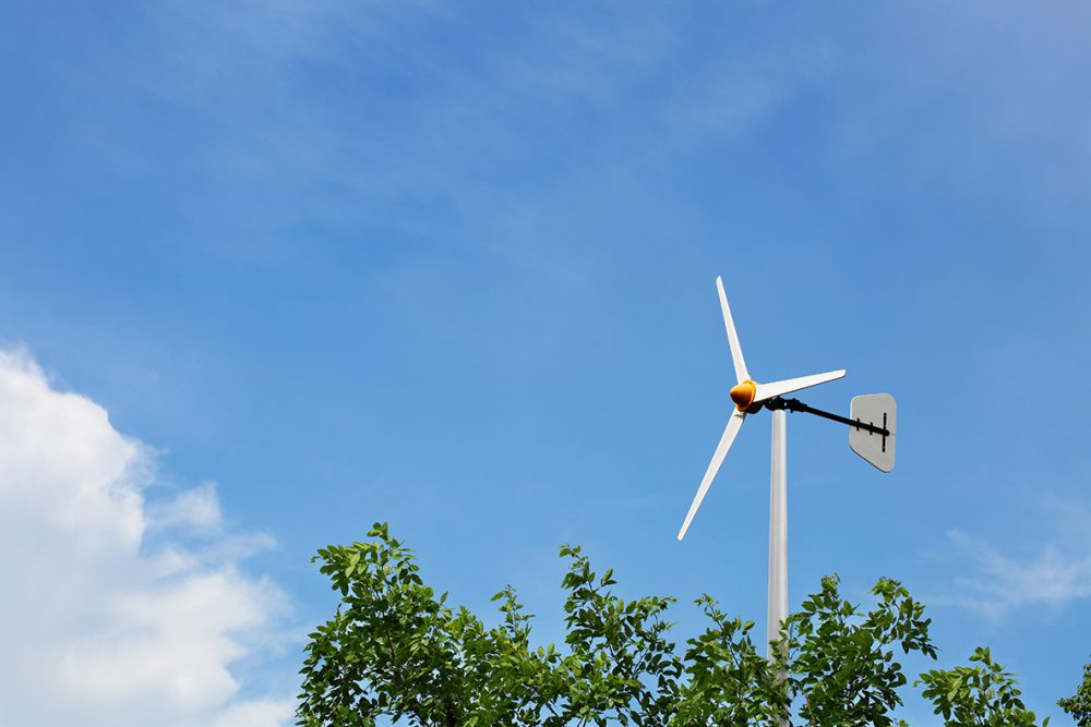 Small white wind turbine with three blades above green trees under clear blue sky